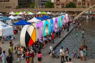 A bustling outdoor event with large, colorful letters spelling 'TORONTO' beside a reflective body of water. Numerous people gather around, some sitting beside the letters, others wading in the water. Around the area, many tents and stalls are set up, creating a lively atmosphere.