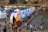 A bustling outdoor event with large, colorful letters spelling 'TORONTO' beside a reflective body of water. Numerous people gather around, some sitting beside the letters, others wading in the water. Around the area, many tents and stalls are set up, creating a lively atmosphere.
