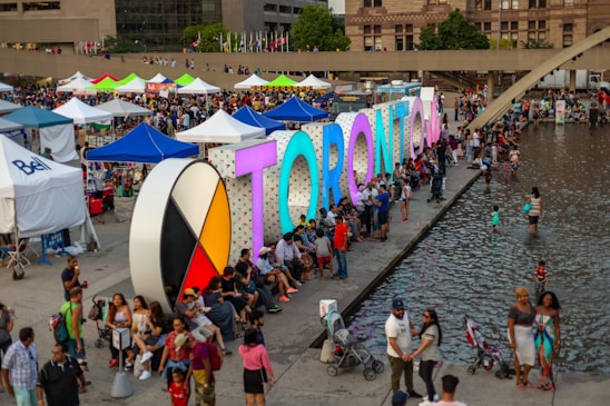 A bustling outdoor event with large, colorful letters spelling 'TORONTO' beside a reflective body of water. Numerous people gather around, some sitting beside the letters, others wading in the water. Around the area, many tents and stalls are set up, creating a lively atmosphere.