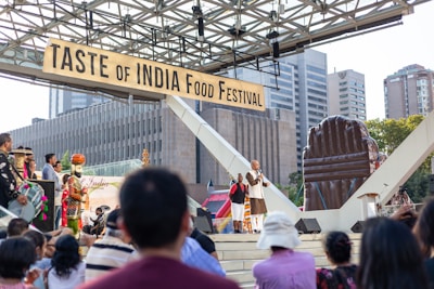 A food festival with a sign reading 'Taste of India Food Festival' takes place outdoors. There is a stage with a person speaking into a microphone, surrounded by attendees. Musical performers with traditional instruments are present. Surrounding the event are modern buildings and an inflatable structure.