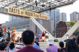 A food festival with a sign reading 'Taste of India Food Festival' takes place outdoors. There is a stage with a person speaking into a microphone, surrounded by attendees. Musical performers with traditional instruments are present. Surrounding the event are modern buildings and an inflatable structure.