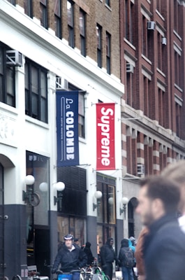 The image depicts a city street scene with several people walking and cycling. Buildings line the street, featuring a 'Supreme' store sign prominently displayed in red and white. Next to it is a 'La Colombe' coffee roasters sign in blue. The atmosphere appears busy with individuals dressed in coats, some wearing helmets. The architecture includes large windows and ornate details typical of urban settings.