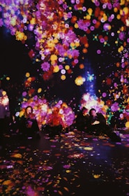 A vibrant display of colorful, luminous flowers is projected onto a dark background, with people sitting amongst the glowing patterns. The surrounding atmosphere is immersive, creating an enchanting and dreamlike ambiance.