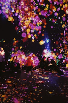 A vibrant display of colorful, luminous flowers is projected onto a dark background, with people sitting amongst the glowing patterns. The surrounding atmosphere is immersive, creating an enchanting and dreamlike ambiance.