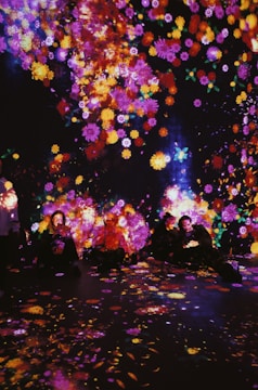 A vibrant display of colorful, luminous flowers is projected onto a dark background, with people sitting amongst the glowing patterns. The surrounding atmosphere is immersive, creating an enchanting and dreamlike ambiance.