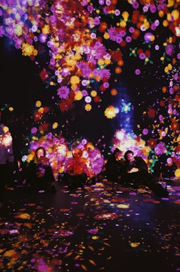 A vibrant display of colorful, luminous flowers is projected onto a dark background, with people sitting amongst the glowing patterns. The surrounding atmosphere is immersive, creating an enchanting and dreamlike ambiance.
