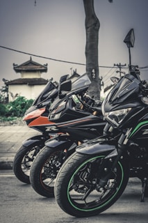 Three sleek motorcycles are lined up closely together on a roadside. The motorcycles are sporty in design, with prominent angular lines and detailed decals. In the background, there is an oriental-style building with a tiered roof, along with a grey sky and some greenery.
