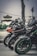 Three sleek motorcycles are lined up closely together on a roadside. The motorcycles are sporty in design, with prominent angular lines and detailed decals. In the background, there is an oriental-style building with a tiered roof, along with a grey sky and some greenery.