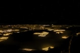 A nighttime scene featuring glowing rectangles scattered across a dark landscape. These illuminated areas resemble greenhouses or fields with artificial lighting. The lights create a soft golden hue against the predominantly black surroundings, with a hint of mist or fog adding an atmospheric effect.