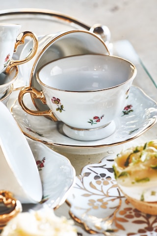 Fine china tea cups and saucers with gold rims, adorned with small floral designs, are artfully arranged. A few delicate pastries sit nearby, partially visible with hints of green garnishes.