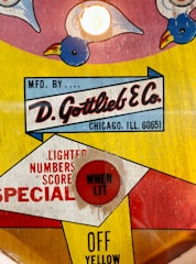 A retro design with colorful graphics featuring a logo for D. Gottlieb & Co. It includes playful illustrations of birds and a red button labeled 'WHEN LIT'. The text also indicates the company's location in Chicago, Illinois. The color palette is a mix of bright yellow, red, and blue.
