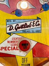 A retro design with colorful graphics featuring a logo for D. Gottlieb & Co. It includes playful illustrations of birds and a red button labeled 'WHEN LIT'. The text also indicates the company's location in Chicago, Illinois. The color palette is a mix of bright yellow, red, and blue.