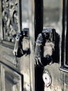 A close-up of an ornate, glossy black door featuring two intricately designed metal knockers shaped like hands. The craftsmanship of the door includes detailed carvings and textures, adding to its antique appearance. A keyhole is also visible below the knockers.