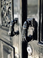 A close-up of an ornate, glossy black door featuring two intricately designed metal knockers shaped like hands. The craftsmanship of the door includes detailed carvings and textures, adding to its antique appearance. A keyhole is also visible below the knockers.