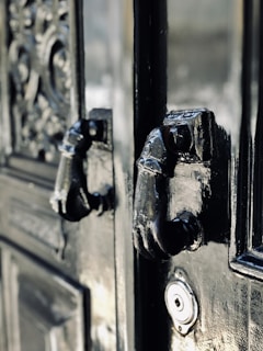 A close-up of an ornate, glossy black door featuring two intricately designed metal knockers shaped like hands. The craftsmanship of the door includes detailed carvings and textures, adding to its antique appearance. A keyhole is also visible below the knockers.