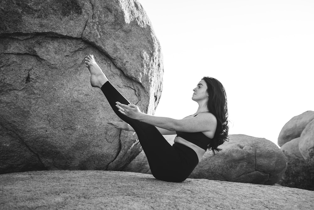 Foto in scala di grigi di una donna che si esercita in cima a una grande roccia