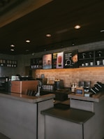 A cozy coffee shop interior featuring a counter with a coffee machine, ingredient dispensers, and signage displaying drink options. The setting includes a brick wall with shelves holding cups and decorative items, under warm, ambient lighting.