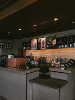 A cozy coffee shop interior featuring a counter with a coffee machine, ingredient dispensers, and signage displaying drink options. The setting includes a brick wall with shelves holding cups and decorative items, under warm, ambient lighting.