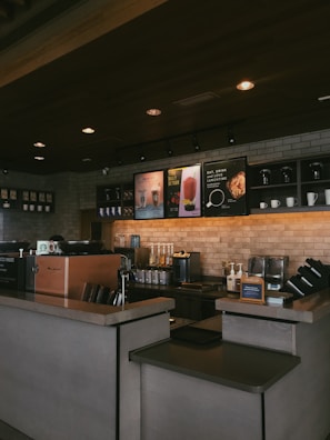A cozy coffee shop interior featuring a counter with a coffee machine, ingredient dispensers, and signage displaying drink options. The setting includes a brick wall with shelves holding cups and decorative items, under warm, ambient lighting.