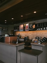 A cozy coffee shop interior featuring a counter with a coffee machine, ingredient dispensers, and signage displaying drink options. The setting includes a brick wall with shelves holding cups and decorative items, under warm, ambient lighting.