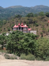 A house with a red roof and multiple gables is nestled on a hillside, surrounded by trees and vegetation. The landscape features rolling hills and a clear blue sky, giving the setting a tranquil and secluded appearance.