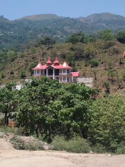 A house with a red roof and multiple gables is nestled on a hillside, surrounded by trees and vegetation. The landscape features rolling hills and a clear blue sky, giving the setting a tranquil and secluded appearance.