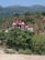 A house with a red roof and multiple gables is nestled on a hillside, surrounded by trees and vegetation. The landscape features rolling hills and a clear blue sky, giving the setting a tranquil and secluded appearance.
