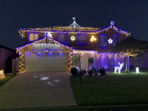 A house is decorated with an elaborate display of Christmas lights, featuring a mix of blue and white colors. There are stars and icicle lights along the roofline, projecting a festive atmosphere. The driveway has snowflake patterns projected onto it. A reindeer light figure is positioned on the lawn, adding to the holiday theme.