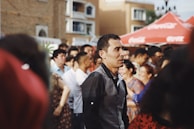 A group of people is gathered outdoors in a social setting. The scene suggests a lively atmosphere, with some individuals engaged in conversation. Red tents are visible in the background with a building facade behind them. The lighting is warm, contributing to the cheerful and vibrant mood.