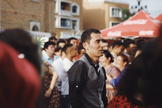 A group of people is gathered outdoors in a social setting. The scene suggests a lively atmosphere, with some individuals engaged in conversation. Red tents are visible in the background with a building facade behind them. The lighting is warm, contributing to the cheerful and vibrant mood.