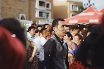 A group of people is gathered outdoors in a social setting. The scene suggests a lively atmosphere, with some individuals engaged in conversation. Red tents are visible in the background with a building facade behind them. The lighting is warm, contributing to the cheerful and vibrant mood.