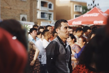 A group of people is gathered outdoors in a social setting. The scene suggests a lively atmosphere, with some individuals engaged in conversation. Red tents are visible in the background with a building facade behind them. The lighting is warm, contributing to the cheerful and vibrant mood.
