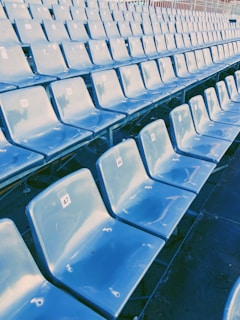 A series of empty blue plastic stadium seats organized in rows, each seat individually numbered with white stickers. The seating area appears to be outdoors, indicated by the metal framework and slight sunlight reflections on the chair surfaces.