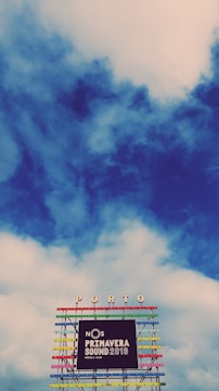 A vibrant and colorful festival sign is displayed against a backdrop of a partly cloudy sky. The structure has multiple horizontal bars in various bright colors, supporting the words 'NOS Primavera Sound 2019' along with the location 'Porto' at the top.