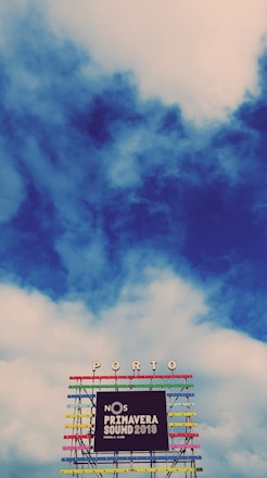 A vibrant and colorful festival sign is displayed against a backdrop of a partly cloudy sky. The structure has multiple horizontal bars in various bright colors, supporting the words 'NOS Primavera Sound 2019' along with the location 'Porto' at the top.