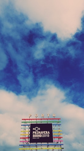A vibrant and colorful festival sign is displayed against a backdrop of a partly cloudy sky. The structure has multiple horizontal bars in various bright colors, supporting the words 'NOS Primavera Sound 2019' along with the location 'Porto' at the top.