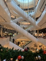 A multi-level shopping mall with several escalators crisscrossing vertically. The area is decorated with strings of small lights and festive greenery, including pine branches adorned with colorful ornaments like red, gold, and silver balls. Numerous shoppers are visible on the escalators and in the background, emphasizing a busy and lively environment.