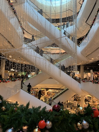 A multi-level shopping mall with several escalators crisscrossing vertically. The area is decorated with strings of small lights and festive greenery, including pine branches adorned with colorful ornaments like red, gold, and silver balls. Numerous shoppers are visible on the escalators and in the background, emphasizing a busy and lively environment.