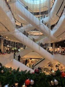 A multi-level shopping mall with several escalators crisscrossing vertically. The area is decorated with strings of small lights and festive greenery, including pine branches adorned with colorful ornaments like red, gold, and silver balls. Numerous shoppers are visible on the escalators and in the background, emphasizing a busy and lively environment.