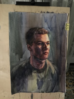 A detailed watercolor portrait of a person with short hair, displayed on an easel. The painting uses dark and muted colors, creating a dramatic and introspective atmosphere.