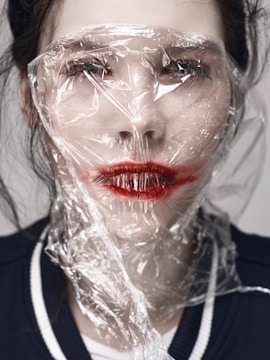 A person with red lipstick is covered with a sheet of transparent plastic material, creating a distorted and unsettling effect over their face. The light hits the crumpled plastic, causing reflections and highlights.
