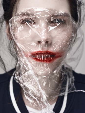 A person with red lipstick is covered with a sheet of transparent plastic material, creating a distorted and unsettling effect over their face. The light hits the crumpled plastic, causing reflections and highlights.