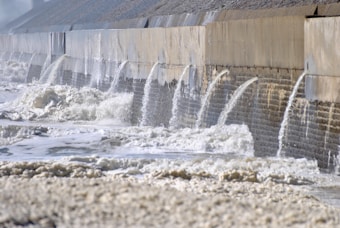 A series of water streams flow through concrete barriers, creating foamy waves in a turbulent body of water. The scene suggests the presence of a dam or spillway handling overflow or runoff.