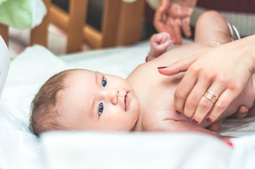 A baby lying on a soft white surface, looking upwards with bright eyes. Gentle hands with manicured nails and rings are tending to the baby, suggesting a nurturing interaction. The background features a crib rail and soft, blurred shapes, contributing to a peaceful and caring setting.
