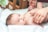 A baby lying on a soft white surface, looking upwards with bright eyes. Gentle hands with manicured nails and rings are tending to the baby, suggesting a nurturing interaction. The background features a crib rail and soft, blurred shapes, contributing to a peaceful and caring setting.