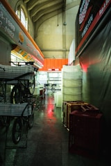 A dimly lit indoor corridor with high ceilings and decorative arches. The left side features a row of delivery bicycles with baskets, while the right side shows stacks of plastic crates. A red glow illuminates parts of the floor and some signage.