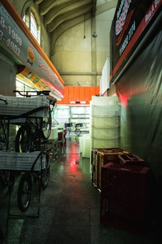 A dimly lit indoor corridor with high ceilings and decorative arches. The left side features a row of delivery bicycles with baskets, while the right side shows stacks of plastic crates. A red glow illuminates parts of the floor and some signage.
