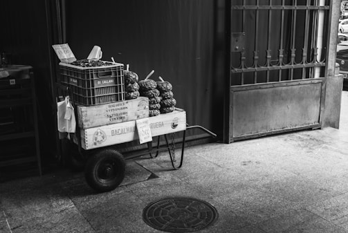 A small cart displays boxes filled with produce, likely chestnuts, against a dark background. The boxes are labeled with various inscriptions, and a handwritten sign indicates a price. The cart is positioned near a gated entrance, creating a rustic market feel.