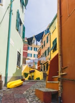 A narrow, picturesque alleyway with colorful buildings featuring pastel hues of green, yellow, orange, and red. The windows have green shutters, and laundry is hanging out to dry between the buildings. The alley is paved with cobblestones, and a bright yellow kayak is placed on the ground. Flower pots and small plants add charm to the area.