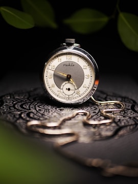 A vintage pocket watch with a silver and white face featuring elegant numerals and intricate detailing rests on an ornate, textured surface. Soft lighting highlights the metallic shine of the watch and its attached chain. Blurred leaves in the background add to the aesthetic composition.